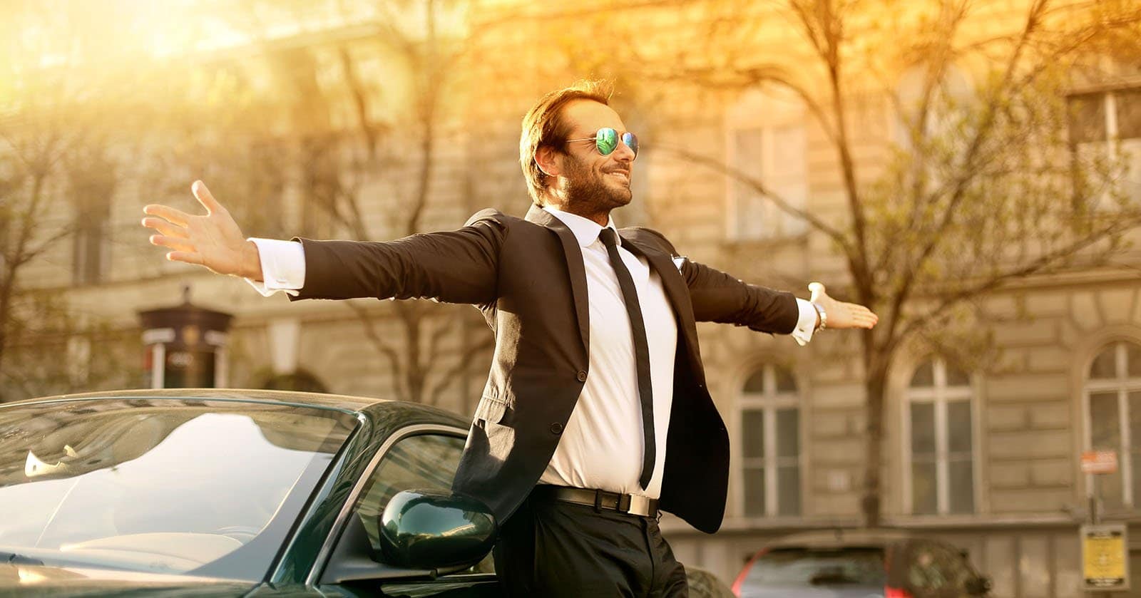 Mann in Anzug und Sonnenbrille steht mit ausgestreckten Armen vor einem Auto